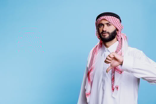 Arab man wearing islamic clothes, showcasing serious expression and giving thumb down portrait. Serious muslim person showing disagreement gesture with finger and looking at camera photo