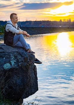 Man Contemplating Nature's Beauty. A man sitting on top of a rock next to a body of water photo