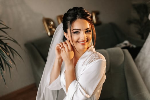 The morning of the bride in a white robe and an elegant hairstyle. An adult girl sits on a chair and fastens an earring, looking into the camera lens and smiling photo