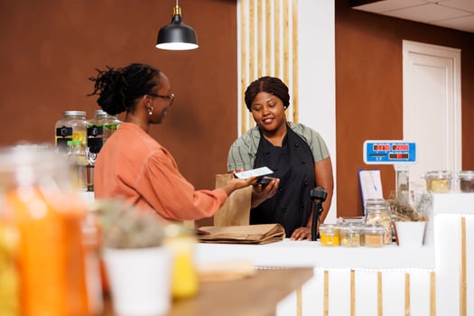Young African American woman, shopping at a modern supermarket, browsing products, and paying with mobile device. The store owner operates the POS terminal at the checkout counter. photo