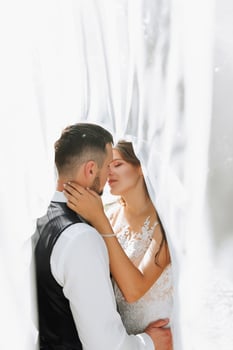 A stylish couple of European newlyweds. Smiling bride in a white dress. The groom, dressed in a classic suit, white shirt, kisses the bride under the veil. Wedding in nature photo