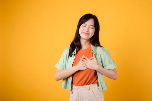 portrait grateful joy young asian woman holding hands to her chest isolated on yellow background. expression peaceful, thankful emotion happiness love positive feeling. photo