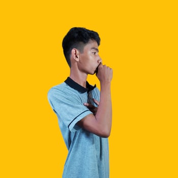 An Asian man is covering his mouth with his hands clenched into fists holding his chest, isolated on a yellow background photo