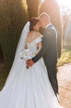 Warm sunny portrait of a happy bride with a bouquet of orchids in her hands and a groom in a classic suit, at sunset. Warm summer time. A long veil, a luxurious white dress with a train. photo