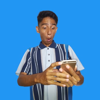 Young asian man surprised looking at smart phone, wearing colorful t-shirt, isolated blue background. photo