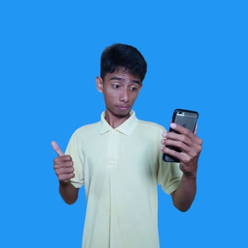 Young Asian man surprised looking at smart phone wearing yellow t-shirt, blue background. photo