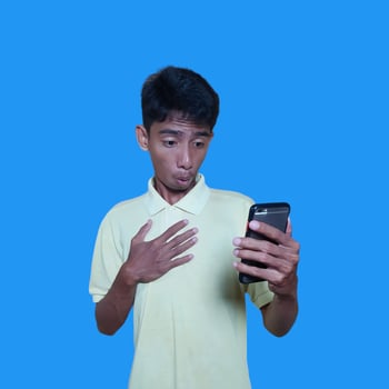 Young Asian man surprised looking at smart phone wearing yellow t-shirt, blue background. photo
