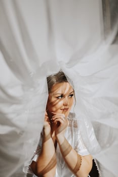 Close-up shot of an elegant brunette bride in a white dress posing under a veil close-up. Bride portrait, professional wedding makeup and hairstyle, bridal fashion. Beautiful bride in a veil photo