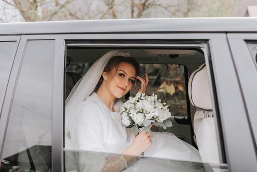 A beautiful smiling bride on her wedding day is sitting in a car and holding a wedding bouquet of white and pink roses. A young woman with a beautiful hairstyle rejoices on her wedding day photo