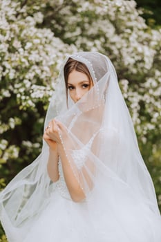 Red-haired bride in a lush dress with an open bust, posing wrapped in a veil, against the background of flowering trees. Spring wedding in nature. photo