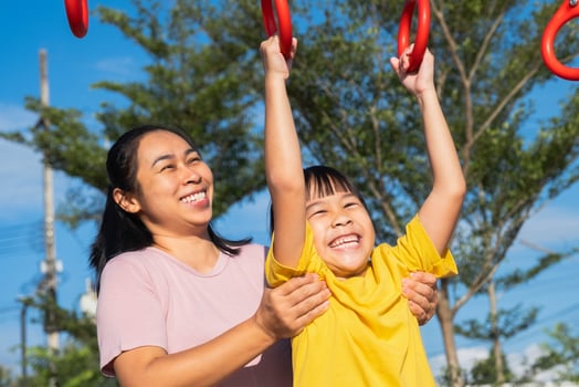 Cute little girl hanging bars in the playground. Happy little Asian child having fun on playground, climbing bars in park. Little girl playing in outdoor playground with mother. photo