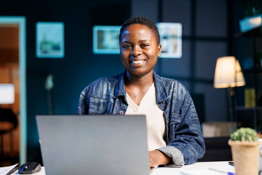 Black woman working at her laptop in a modern apartment, studying online and researching information for her project. Smiling african american blogger using personal computer while looking at camera. photo