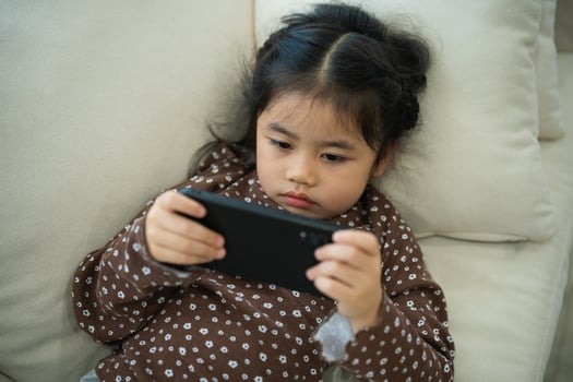 Asian child girl serious or concentrate and using mobile phone screen on couch sofa. Baby smiling funny time use mobile phone. Too much screen time. Girl watching videos while tv, Internet addiction. photo