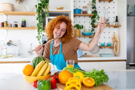 Funny beautiful woman singing into spatula, cooking in modern kitchen, holding spatula as microphone, dancing, listening to music, playful girl having fun with kitchenware, preparing food. photo
