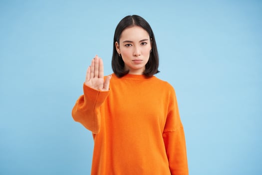 Stop. Serious and confident korean female model, extends hand, shows taboo, rejection gesture, refuse smth, stands over blue background photo