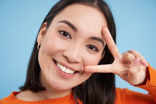 Positive and happy japanese woman, shows peace sign and smiles, happy v-sign gesture, standing over blue background photo