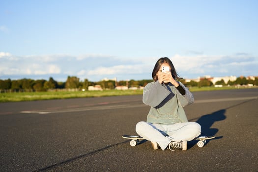Hipster teen girl sitting on her skateboard, taking photos on smartphone. Asian woman skater sits on longboard and photographing on mobile phone