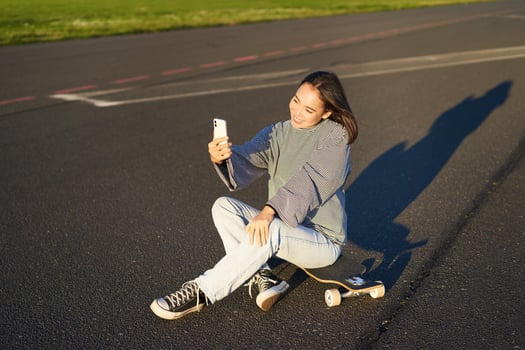 Beautiful korean girl takes selfie on smartphone, takes photo with her skateboard, enoys sunny day outdoors