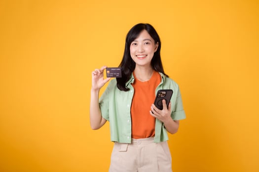 thrill of shopping from mobile with a captivating young Asian woman in her 30s, elegantly dressed in orange shirt and green jumper, using smartphone to show credit card on yellow studio background. photo