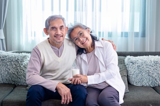 Couple of healthy marriage Asian senior father and mother sitting together with glass of milk and happy smile on cozy couch in retirement home during winter for elder care to spending valuable time photo