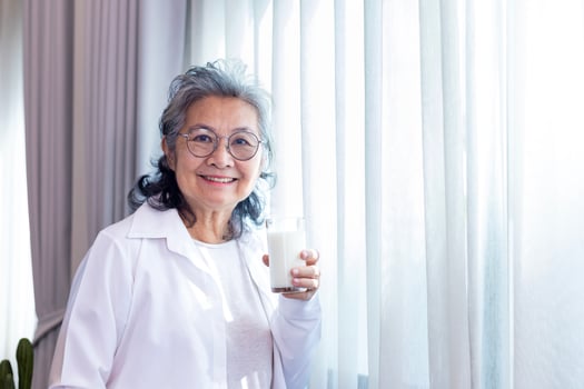 Senior Asian lady drinking glass of milk while standing by the window with copy space for calcium boosting and healthy dairy product consumption concept photo