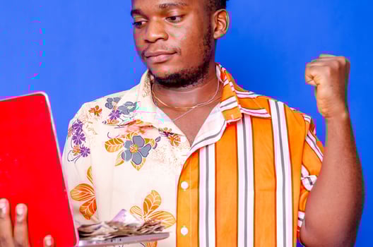 Photo of a happy young afro american handsome man posing isolated over blue wall background using laptop computer holding money.