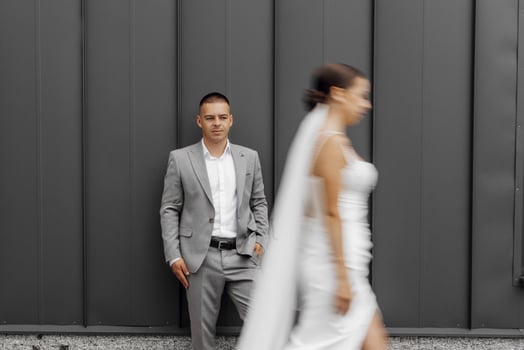 Fashionable and staged photo of the bride and groom against the background of a gray wall. A bride with a long veil blowing in the wind. The bridegroom is looking at the camera