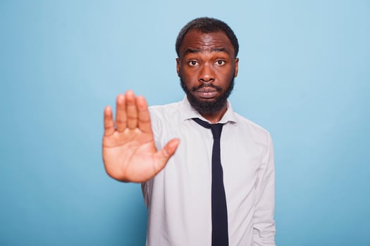 On isolated blue background, serious looking businessman in a white shirt and black tie, makes a hand stop in disapproval. Professional office worker with a grumpy demeanor putting up denial sign. photo
