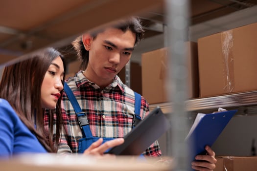 Storehouse asian workers comparing inventory data reports in storage room. Postal warehouse employees doing logistics and distribution management together near cardboard boxes shelf photo