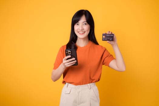 Vibrant young Asian woman in her 30s, using smartphone and holding credit card on yellow background. Convenient online shopping. photo