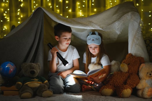 Two children with flashlight read a book under a blanket as a tent. photo