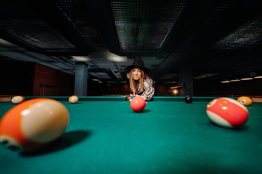 A girl in a hat in a billiard club with a cue in her hands hits a ball.Playing pool photo