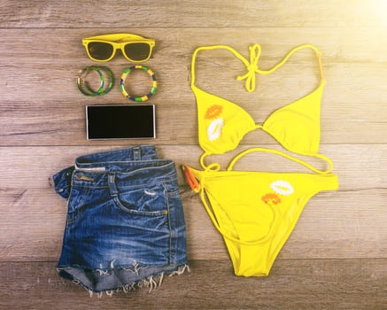 Set of beach clothes yellow bikini, bracelets, jeans shorts, glasses on dark wooden background. Top view. Summer Holiday Concept. photo