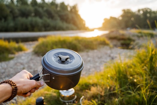 Meal in nature. Pot -pot in the hands of a tourist, focus on them. Ahead of the river landscape and green forest. Nearby lies the rest of the kitchen camping utensils. Tourist snack, outdoor activiti photo