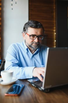 Positive senior bearded man with grey hair drinking coffee and using laptop at cafe, copy space. Stylish aged businessman in burgundy jacket enjoying his tea while working online. photo