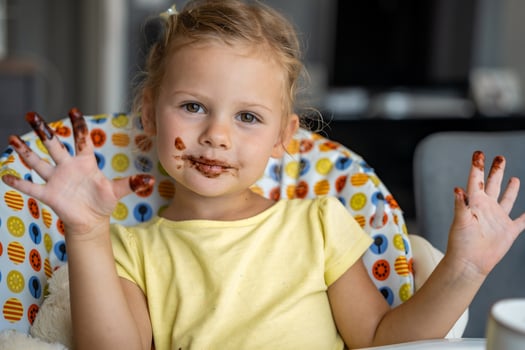 Little girl with blond hair eating homemade chocolate and showing mouth and dirty hands with stains of chocolate in home kitchen photo