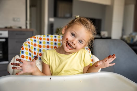 Little girl with blond hair eating homemade chocolate and showing mouth and dirty hands with stains of chocolate in home kitchen photo