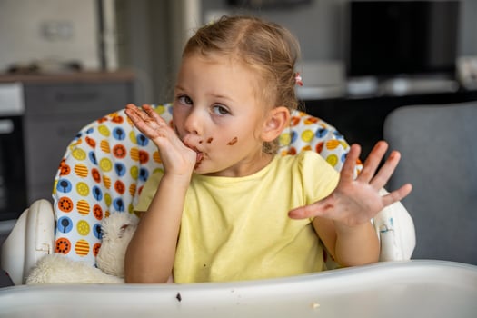 Little girl with blond hair eating homemade chocolate and showing mouth and dirty hands with stains of chocolate in home kitchen photo