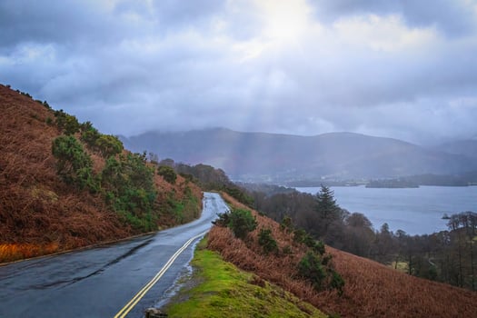 Winding road through a misty landscape with sun rays piercing the clouds over a serene lake and hills. photo