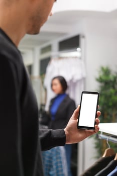 Clothing store customer holding smartphone with blank screen mockup while exploring apparel online. Boutique man hand using mobile phone with empty white display while shopping in mall photo