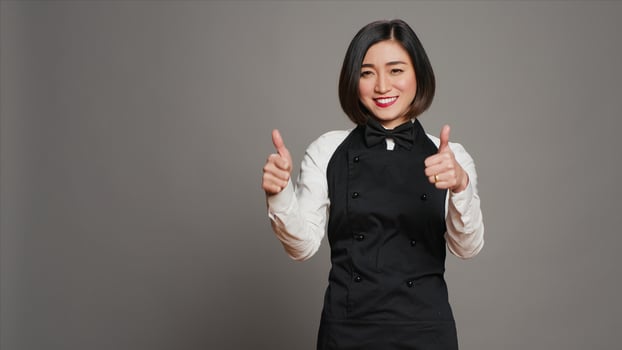 Smiling waitress giving thumbs up sign in front of camera, working in a five star restaurant to serve tables. Asian server with apron showing approval with okay symbol in studio, agreement. Camera A. photo