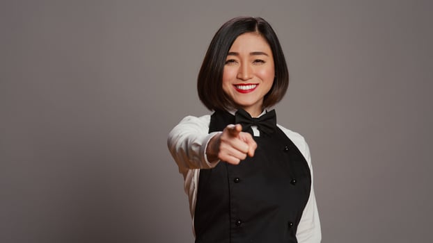 Asian waitress pointing at camera to choose you over grey background, wearing uniform with bow and apron. Woman catering worker choosing someone to work at a restaurant. Camera B. photo