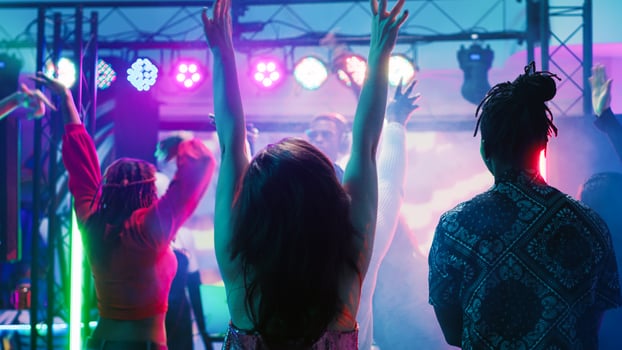 Diverse people partying at nightclub, enjoying live music performance from DJ station at discotheque stage. Group of friends dancing together on dance floor with spotlights. Tripod shot. photo