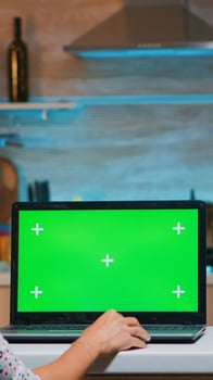 Business woman looking at green screen laptop sitting at home in kitchen. Freelancer watching desktop monitor display with green mockup, chroma key, during night time working overtime. photo