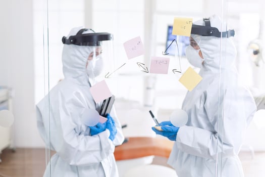 Tired dentist discussing about patient diagnosis wearing ppe suit. Medical team in stomatology office wearing coverall in dental office writing ideas on sticky notes. photo