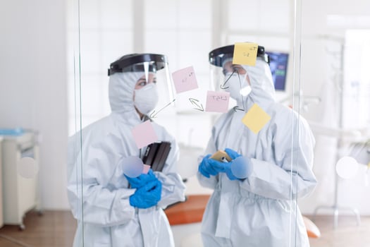 Dentist coworkers talking about teeth issues wearing pee suit as safety precauiton for covid-19. Medical team in stomatology office wearing coverall in dental office writing ideas on sticky notes. photo