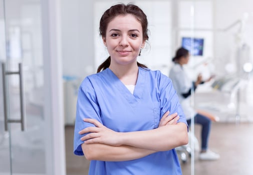 Portrait of nurse in dental reception with arms crossed looking at camera wearing uniform and dentist in the background.. Happy smiling assistant in orthodontic office. photo