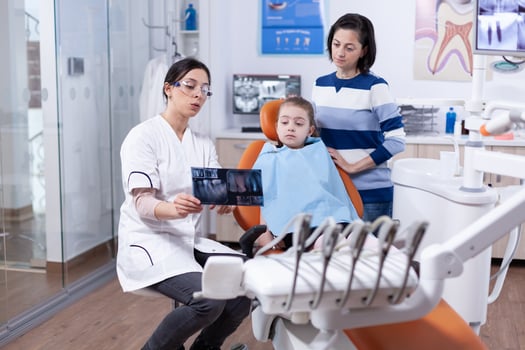 Cheerful little girl sitting in dental office while doctor showing something on radiography. Stomatologist explaining teeth diagnosis to mother of child in health clinic holding x-ray. photo