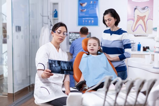 Dentist examining little kid radiography sitting on chair wearing dental bib. Stomatologist explaining teeth diagnosis to mother of child in health clinic holding x-ray. photo