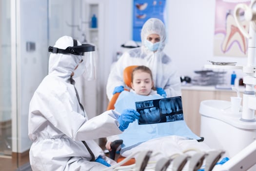 Child and mother in ppe suit listening dentist talking about dental procedure. Stomatolog in protectie suit for coroanvirus as safety precaution holding child teeth x-ray during consultation. photo
